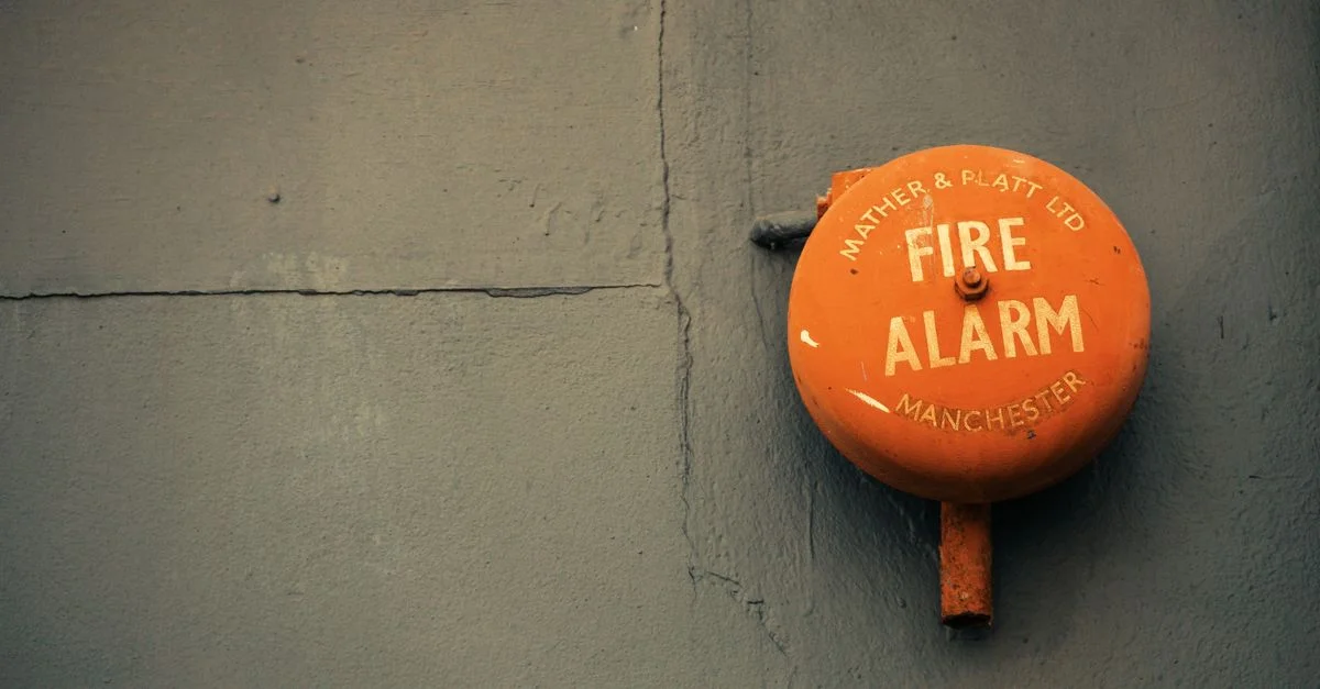 Gros plan d'une vieille alarme incendie sur un mur en béton gris, avec le texte 'Mather & Platt Ltd Alarme Incendie Manchester.'